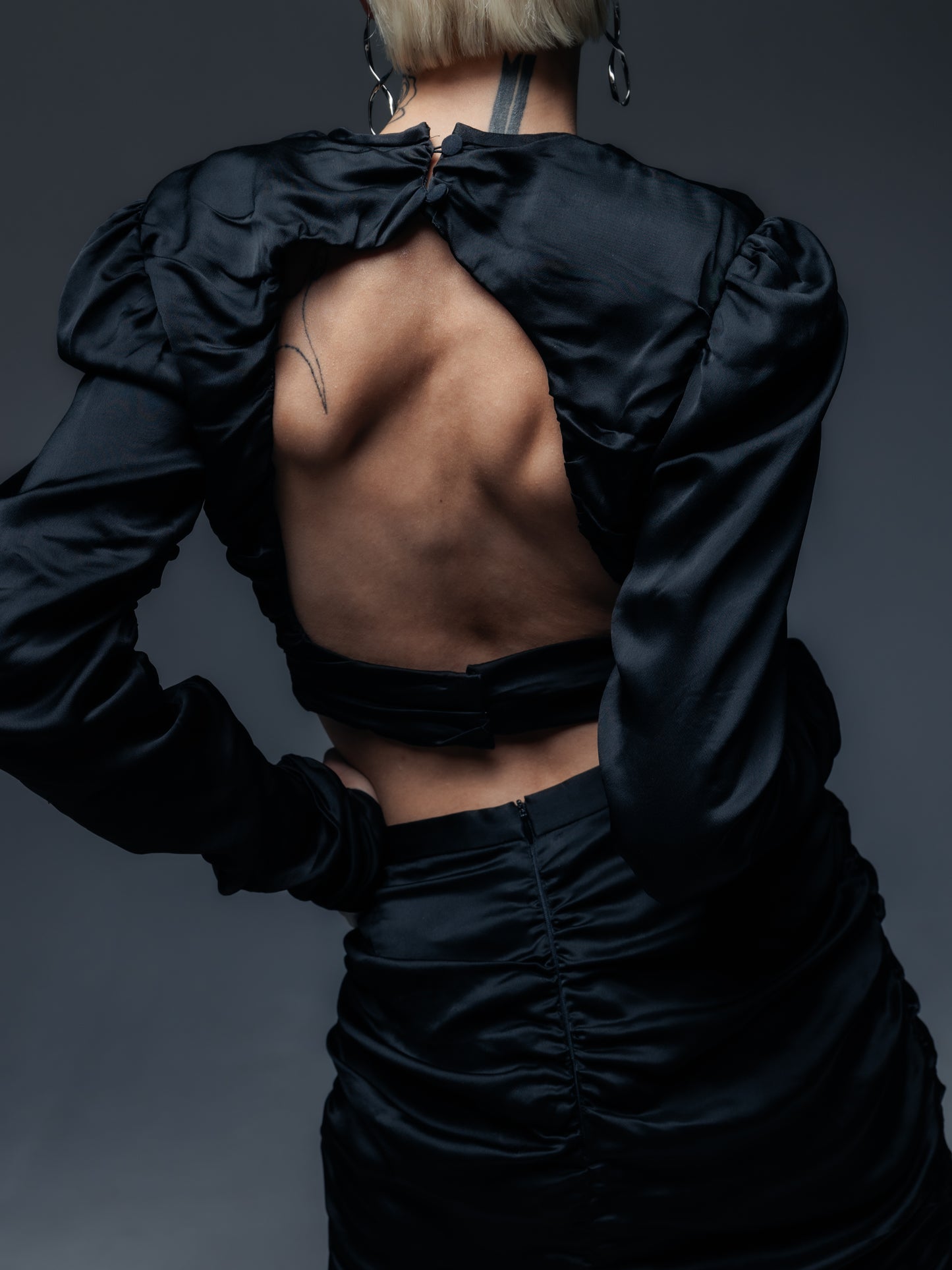 Tylny widok czarnej marszczonej bluzki Jupiter z odkrytymi plecami. Widzoczne wycięcie na plecach podkreśla oryginalny fason bluzki, a dopasowany krój ukazuje subtelny zamek z tyłu spódnicy. Bluzka z długimi, przylegającymi rękawami i marszczeniami prezentuje się zjawiskowo w wieczorowej stylizacji.