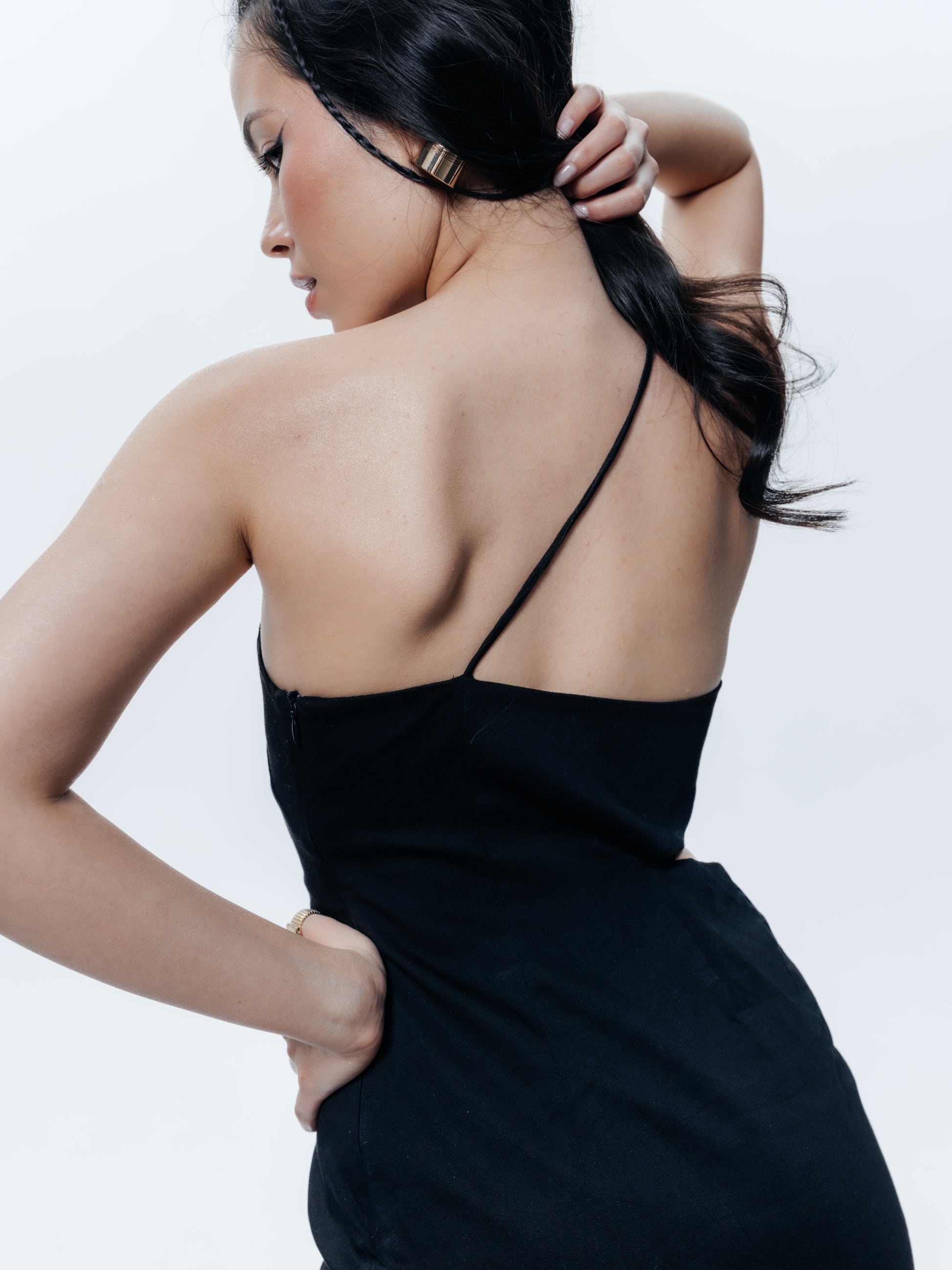 Widok sukienki Mercury z tyłu i boku. Sukienka układa się gładko na plecach – widać, że zapięcie znajduje się na lewym boku (tył jest jednolity, bez zakłóceń). Cienkie ramiączko wychodzące z przodu biegnie przez szyję i łączy się z tylną krawędzią sukienki po prawej stronie, dzięki czemu plecy w dużej części pozostają odkryte. Dopasowany tył podkreśla linię pleców i pośladków, a brak ramiączka na lewej stronie tworzy ciekawą, asymetryczną linię na łopatkach. 