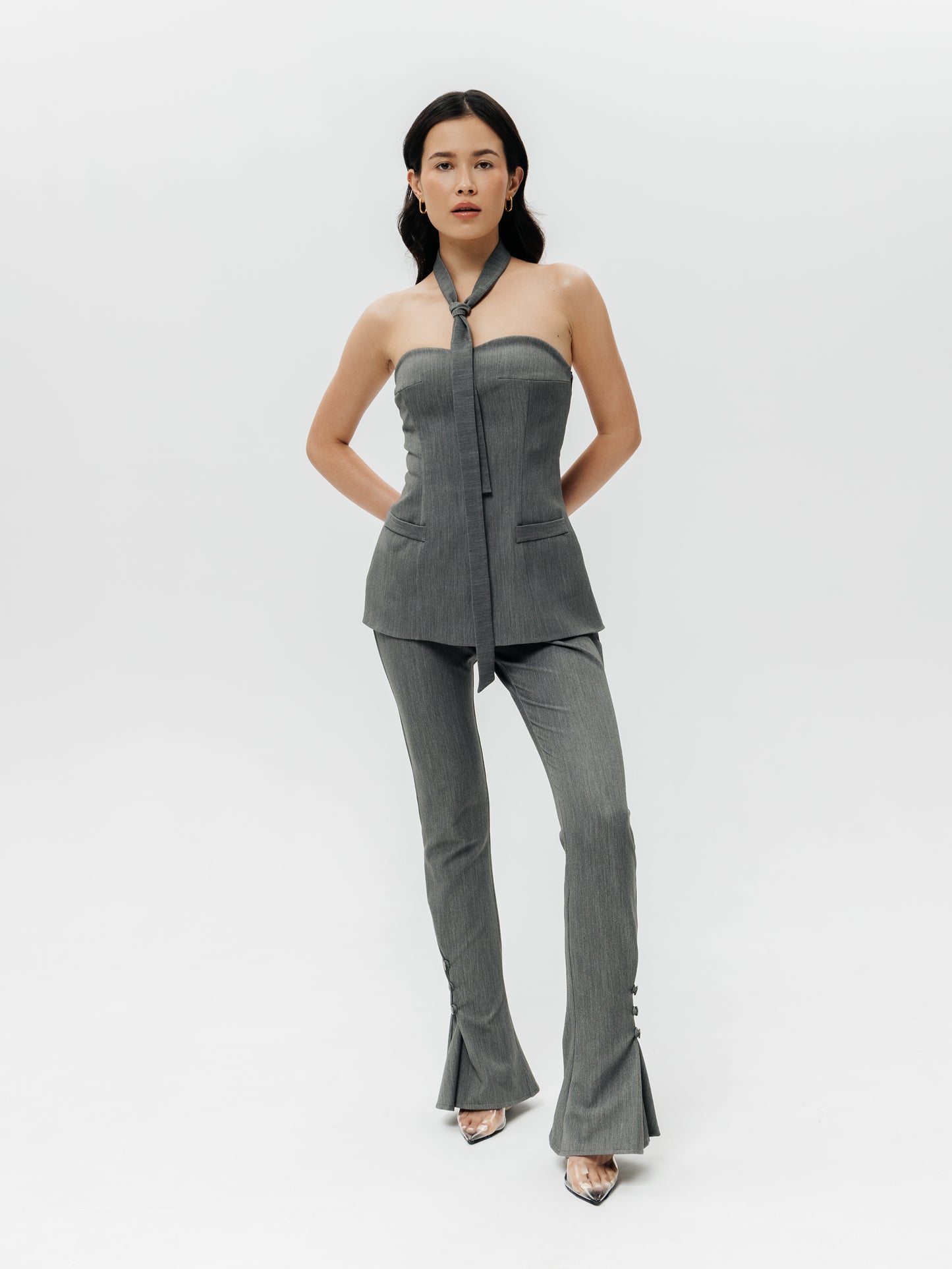 Modelka w dopasowanych szarych spodniach garniturowych Mars, wykonanych z wysokogatunkowej tkaniny o domieszce wiskozy i elastanu. Spodnie mają wysoki stan i unikalny detal – po obu bokach talii znajdują się symetryczne wycięcia ozdobione po dwoma czarnymi guzikami, które dodają wyrafinowania. Nogawki są proste, z wszytymi godetami na dole, które tworzą subtelne rozcięcia; każde rozcięcie zapinane jest na trzy małe guziki, co nadaje spodniom oryginalny akcent. 