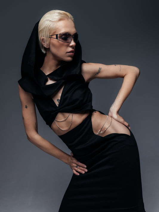 Dynamiczne ujęcie sukienki Moon z profilu: modelka stoi bokiem z ręką opartą na biodrze, co uwydatnia detale kreacji. Z tej perspektywy wyraźnie widać, jak srebrne łańcuszki zdobią zarówno odsłonięty przód sukienki, jak i wycięcia na biodrach – układają się one w efektowne, połyskujące linie wzdłuż ciała. Czerń miękkiej, bawełnianej tkaniny kontrastuje z metalicznym blaskiem łańcuszków, podkreślając śmiały design projektu. Kaptur okalający głowę modelki oraz futurystyczne dodatki, jak np. okulary.