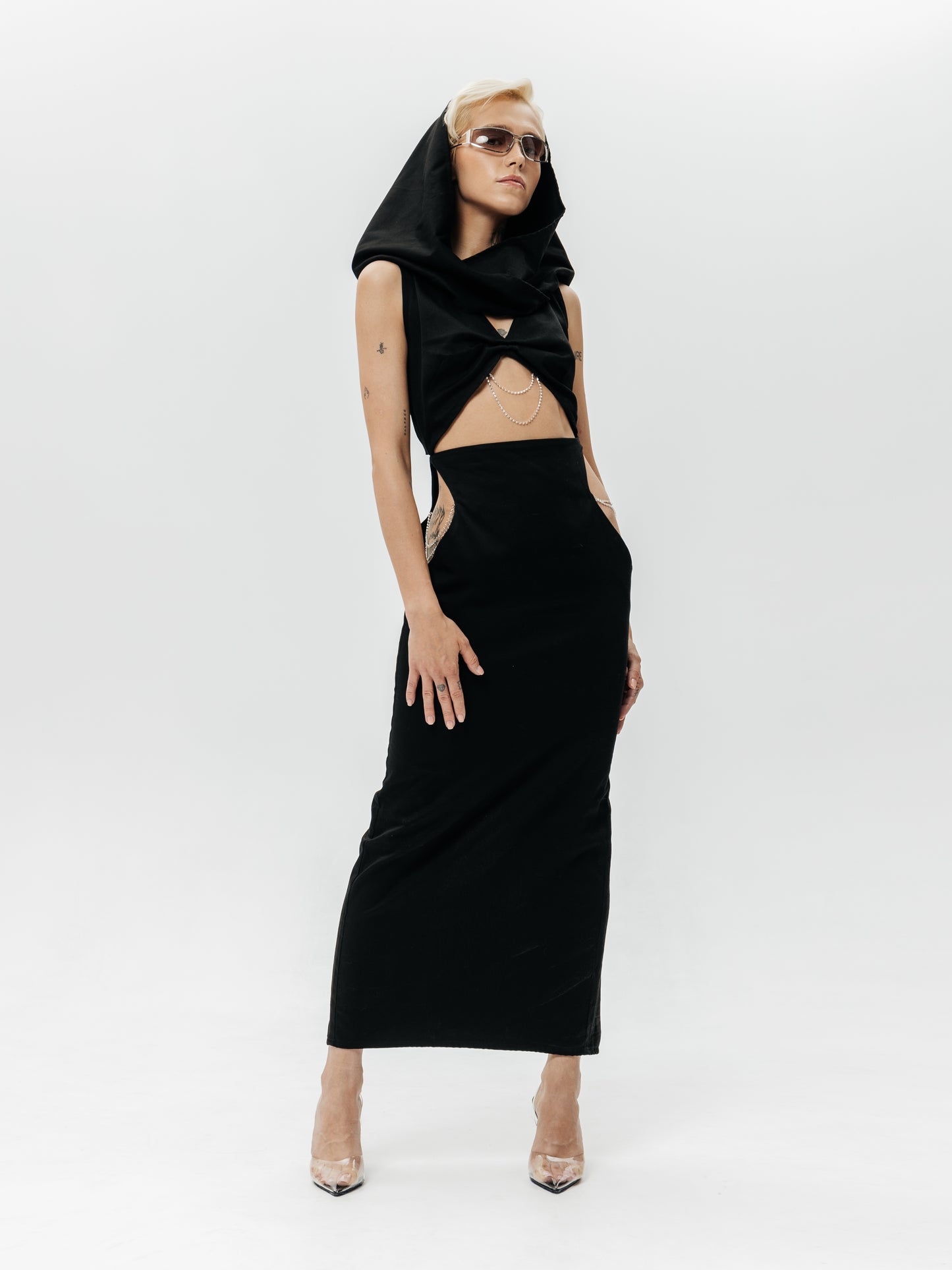 Modelka w czarnej sukience Moon z kapturem, ukazana od przodu, prezentuje kreację w pełnej okazałości. Dopasowany, długi fason bez rękawów sięga do kostek, podkreślając smukłą sylwetkę. Na wysokości talii i brzucha widoczne jest zmysłowe wycięcie ozdobione subtelnym srebrnym łańcuszkiem, co dodaje kreacji drapieżnego charakteru. Po bokach bioder sukienki połyskują srebrne łańcuszki w wąskich wycięciach, przyciągając wzrok do talii. Kaptur założony na głowę nadaje stylizacji nutę tajemnicy i niepowtar