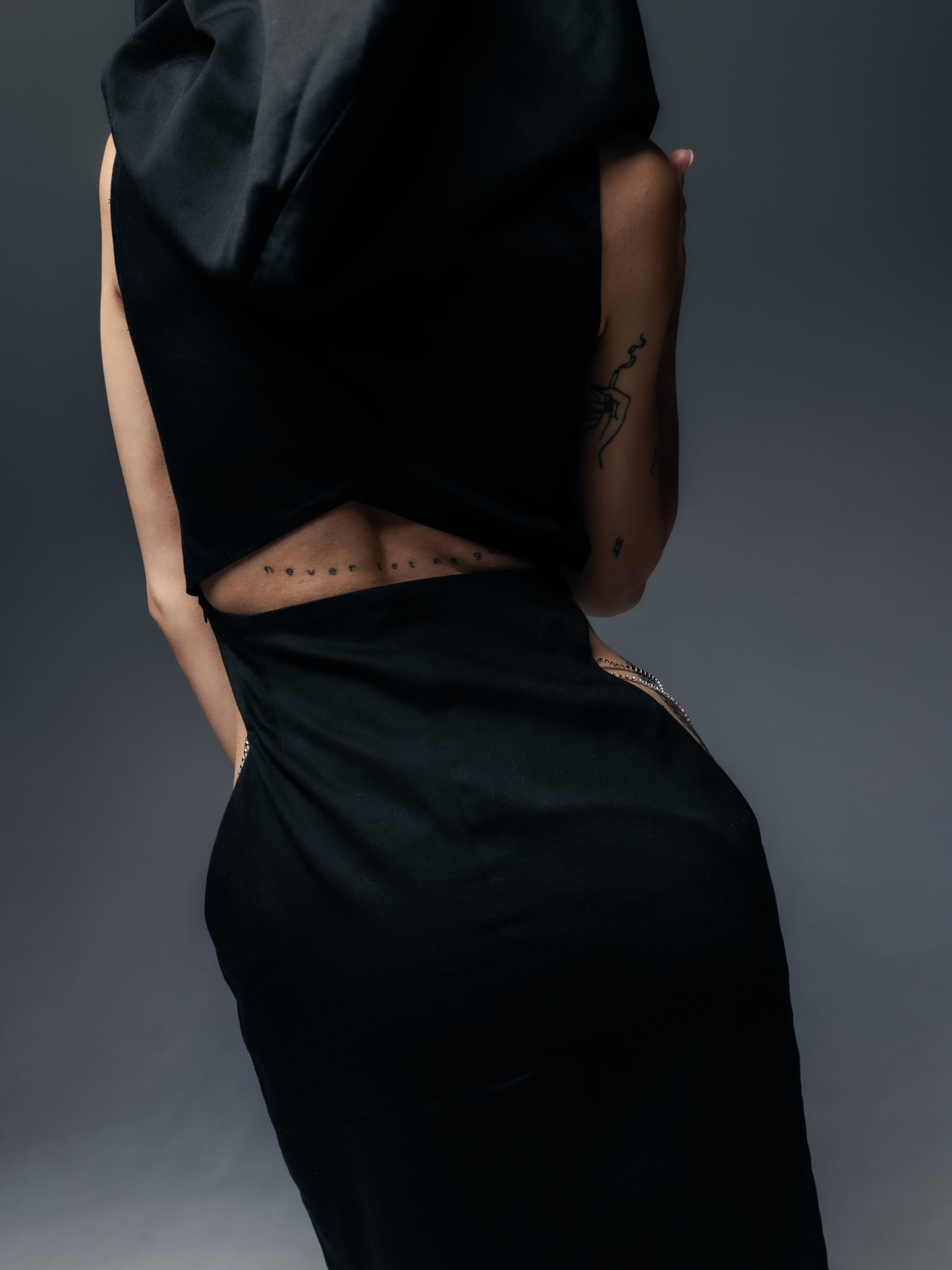 Tylny widok sukienki Moon ukazuje odsłonięte plecy i biodra modelki. Głębokie wycięcie na plecach eksponuje skórę w okolicy talii, nadając kreacji odważny charakter. Obszerny kaptur luźno opada na ramiona z tyłu, zachowując swoją formę i dodając sylwetce enigmatycznego uroku nawet z tej perspektywy. Po obu stronach talii widać symetryczne wycięcia ozdobione srebrnymi łańcuszkami, które błyszczą przy ruchu, podkreślając krągłość bioder. 