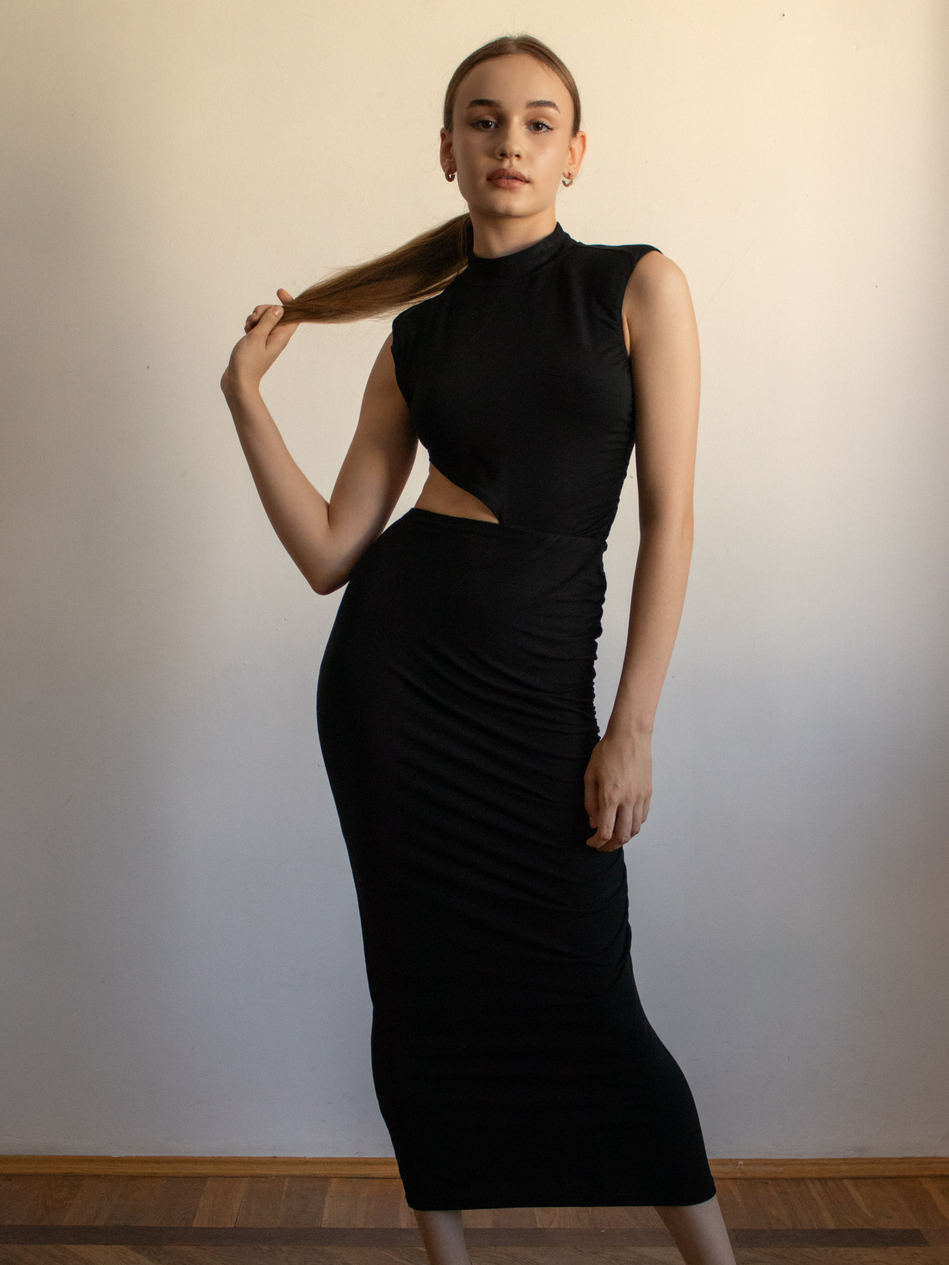 Długa, czarna sukienka mocno przylega do ciała, ukazując linię bioder i ud, a marszczenie materiału po prawej stronie dodaje tekstury. Wycięcie w talii przyciąga wzrok do smukłej figury modelki. Stójka przy szyi nadaje kreacji nieco futurystyczny, modowy charakter. To pełne ujęcie sylwetki pokazuje, jak sukienka Uranus łączy skromność formy (długi, zabudowany fason) z odważnym elementem (asymetryczne wycięcie), tworząc efektowną kreację na wieczór w stylu miejskiej elegancji.