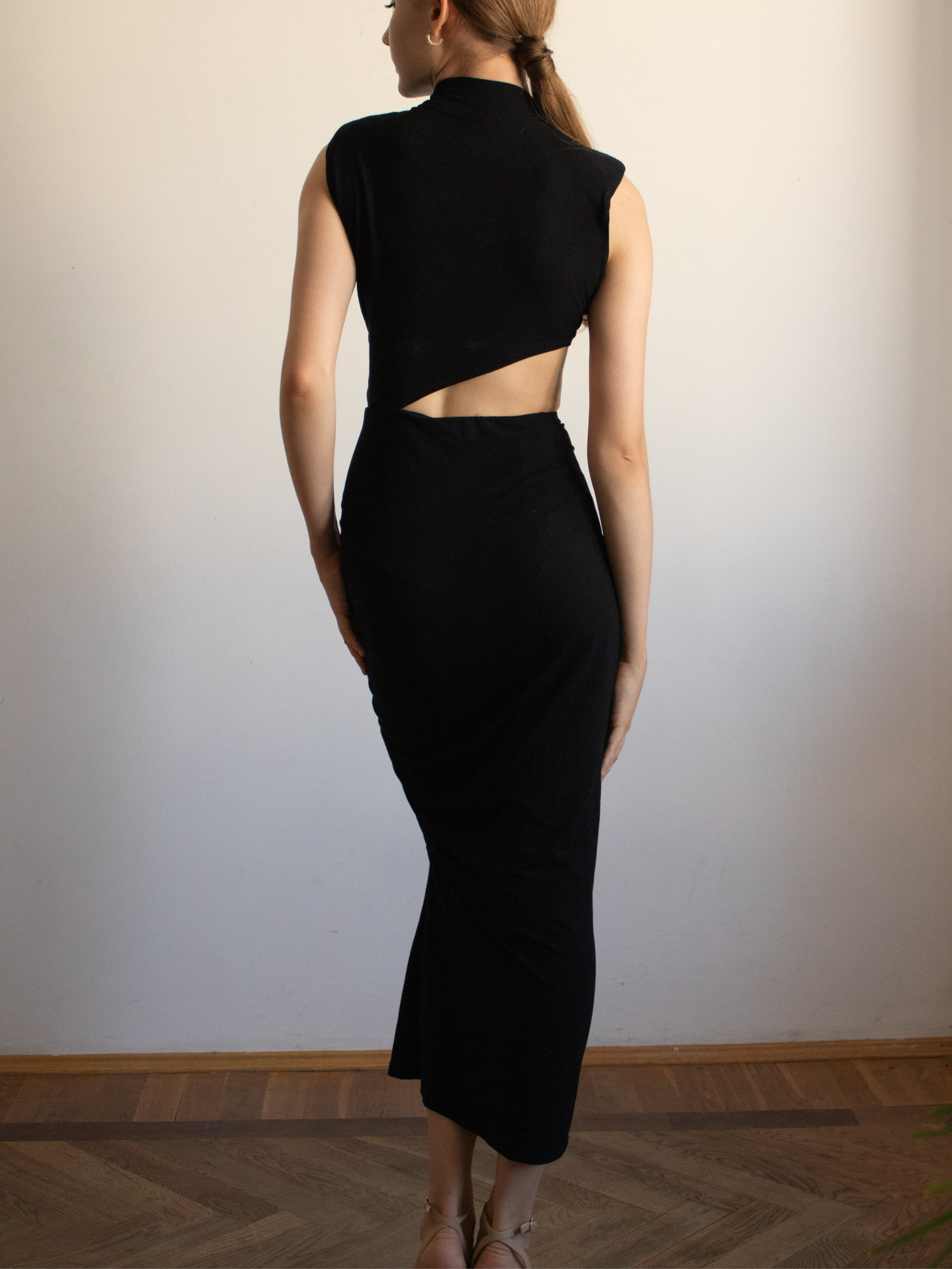 Sylwetka z tyłu sukienki jest prosta i gładka. Asymetryczne wycięcie z boku układa się również na plecach, tworząc ciekawą linię – lewa strona pleców i talii jest częściowo odsłonięta, podczas gdy prawa pozostaje zakryta aż po łopatkę. Sukienka nie posiada tradycyjnych ramiączek ani zapięć, co potęguje minimalistyczny charakter tyłu. Ten widok podkreśla zgrabną figurę i odważny krój projektu, ale nadal pozostaje elegancki dzięki długości midi i stonowanej formie.