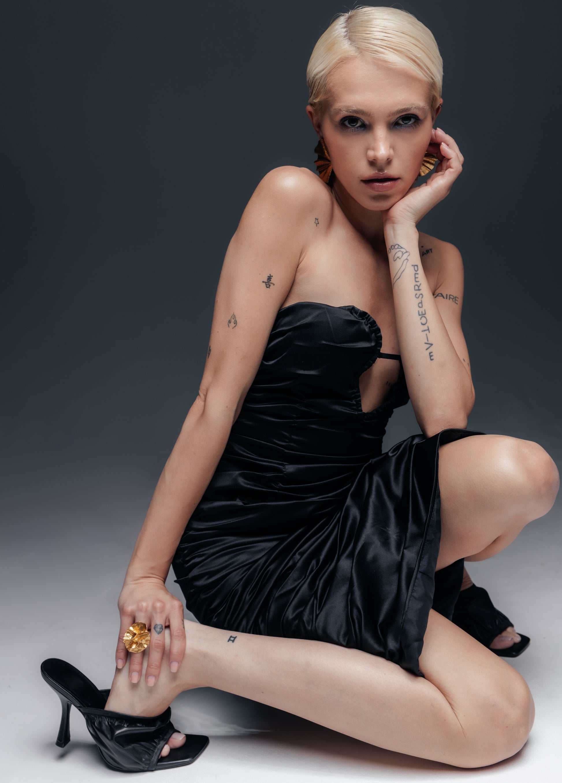 czarna satyna miękko układa się przy ruchu – marszczenia przemieszczają się, tworząc za każdym razem niepowtarzalny wzór drapowania. Odsłonięte ramiona i obojczyki dodają zmysłowości, a długość midi podkreśla smukłość kostek i elegancję obuwia. Sukienka Venus w takiej artystycznej odsłonie pokazuje swój charakter: to kreacja dla pewnej siebie kobiety, która ceni klasyczną czerń, lecz pragnie również oryginalnego detalu i efektu „wow” w stylizacji.