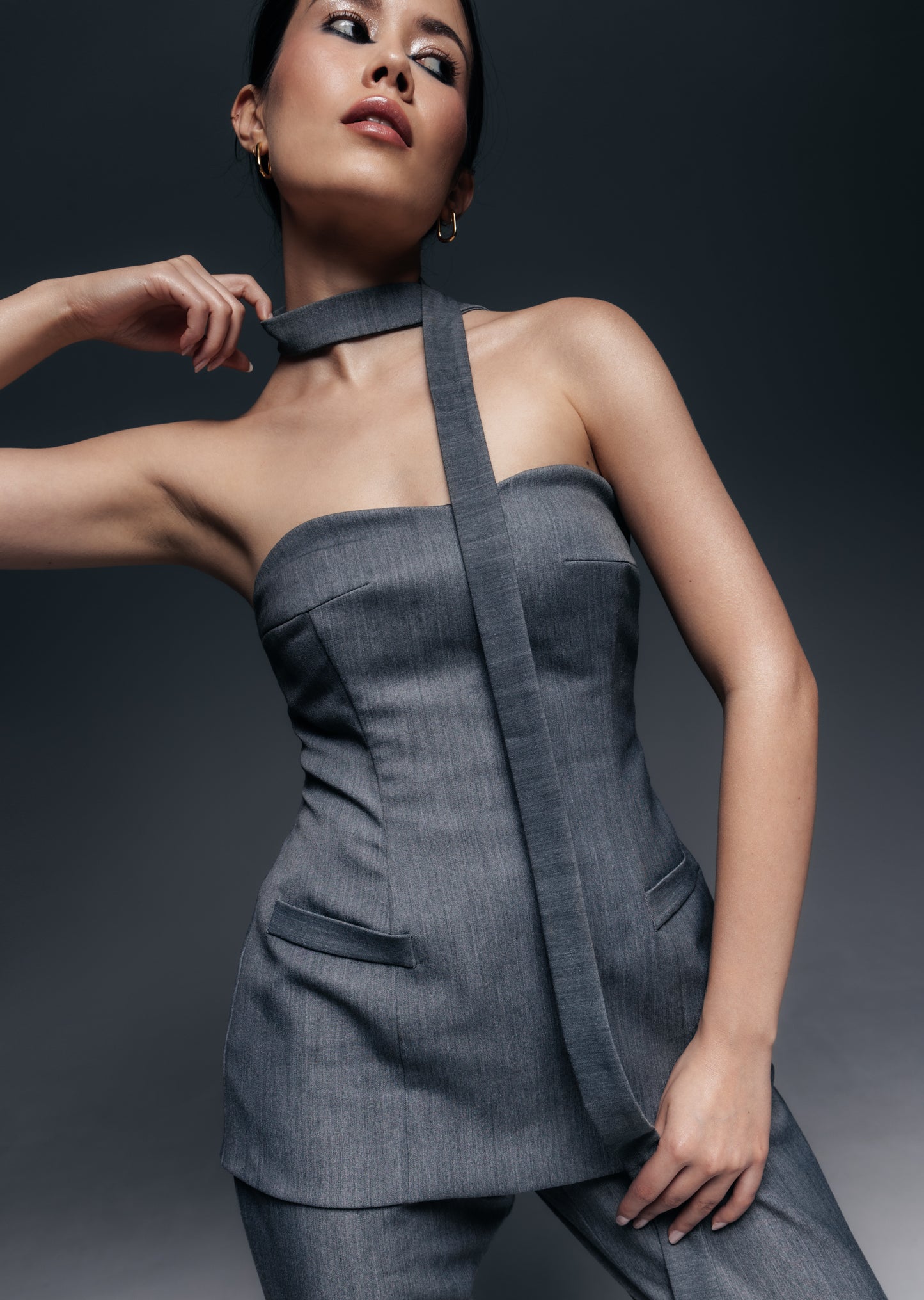  Długi, wąski szal Mars w kolorze szarego melanżu, noszony jako dodatek do garniturowego kompletu. Szal ma około 145 cm długości i jedynie 3 cm szerokości – to cienka, elegancka tasiemka z tej samej garniturowej tkaniny co spodnie i gorset Mars (poliester z wiskozą). Modelka przewiązała szal wokół szyi niczym krawat lub choker – jeden koniec zawiązany jest z przodu na prosty węzeł, a drugi zwisa swobodnie, sięgając okolic talii. Taki sposób noszenia podkreśla smukłość szyi i dodaje modowego sznytu.