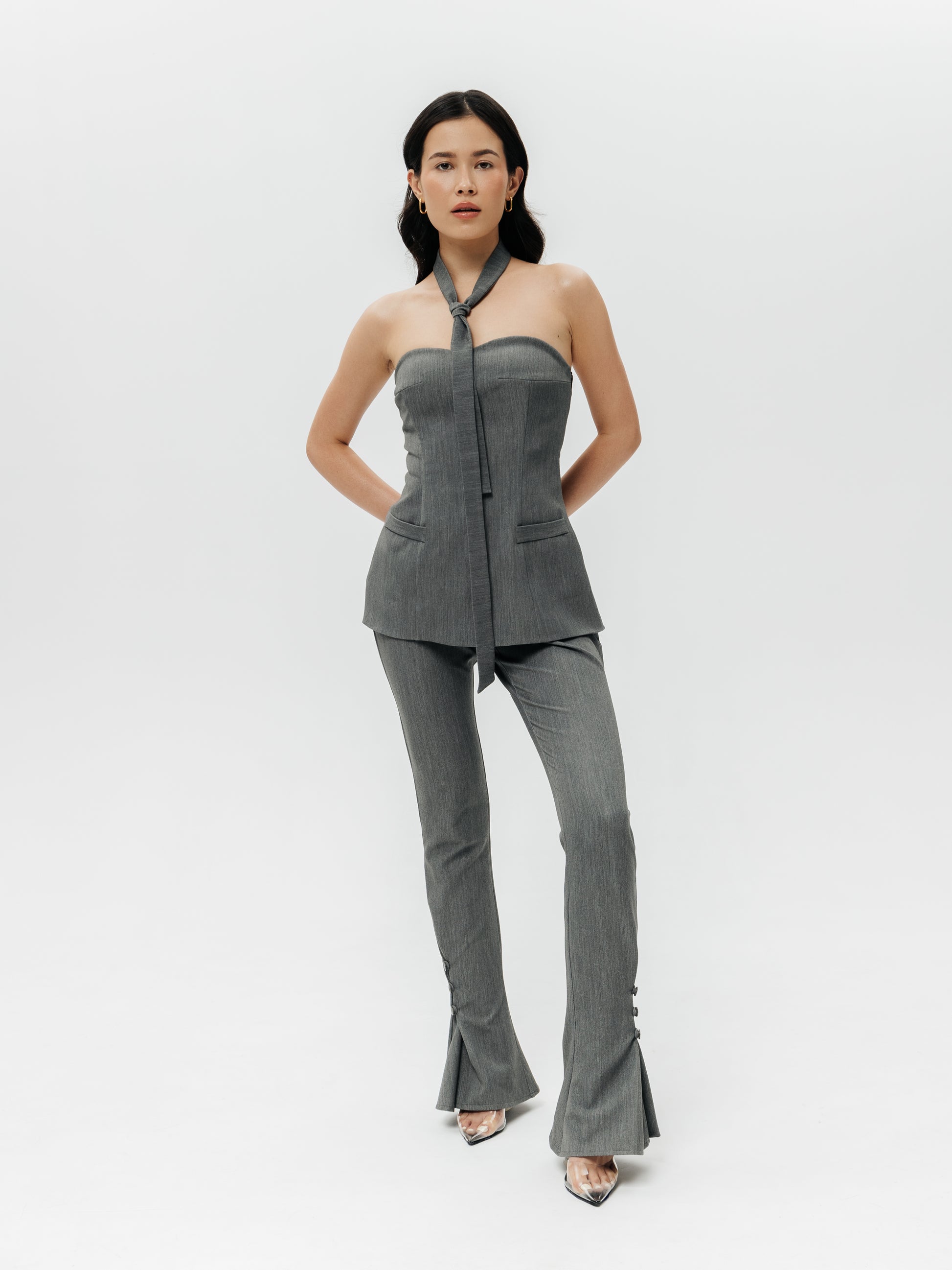 Pełne ujęcie garniturowego zestawu z topem Mars na jasnym tle. Modelka ma na sobie gorset Mars bez ramiączek, który idealnie przylega na biuście (dzięki fiszbinom tworzy zgrabną linię dekoltu) i luźniej opływa biodra, tworząc modny, wydłużony kształt. Do topu dobrane są szare spodnie Mars z wysokim stanem i rozszerzanymi, rozciętymi dołami – razem tworzy to nietuzinkowy damski garnitur wieczorowy. Cienki szal Mars zawiązany na kształt krawata dopełnia look, nadając mu odrobinę nonszalancji. 