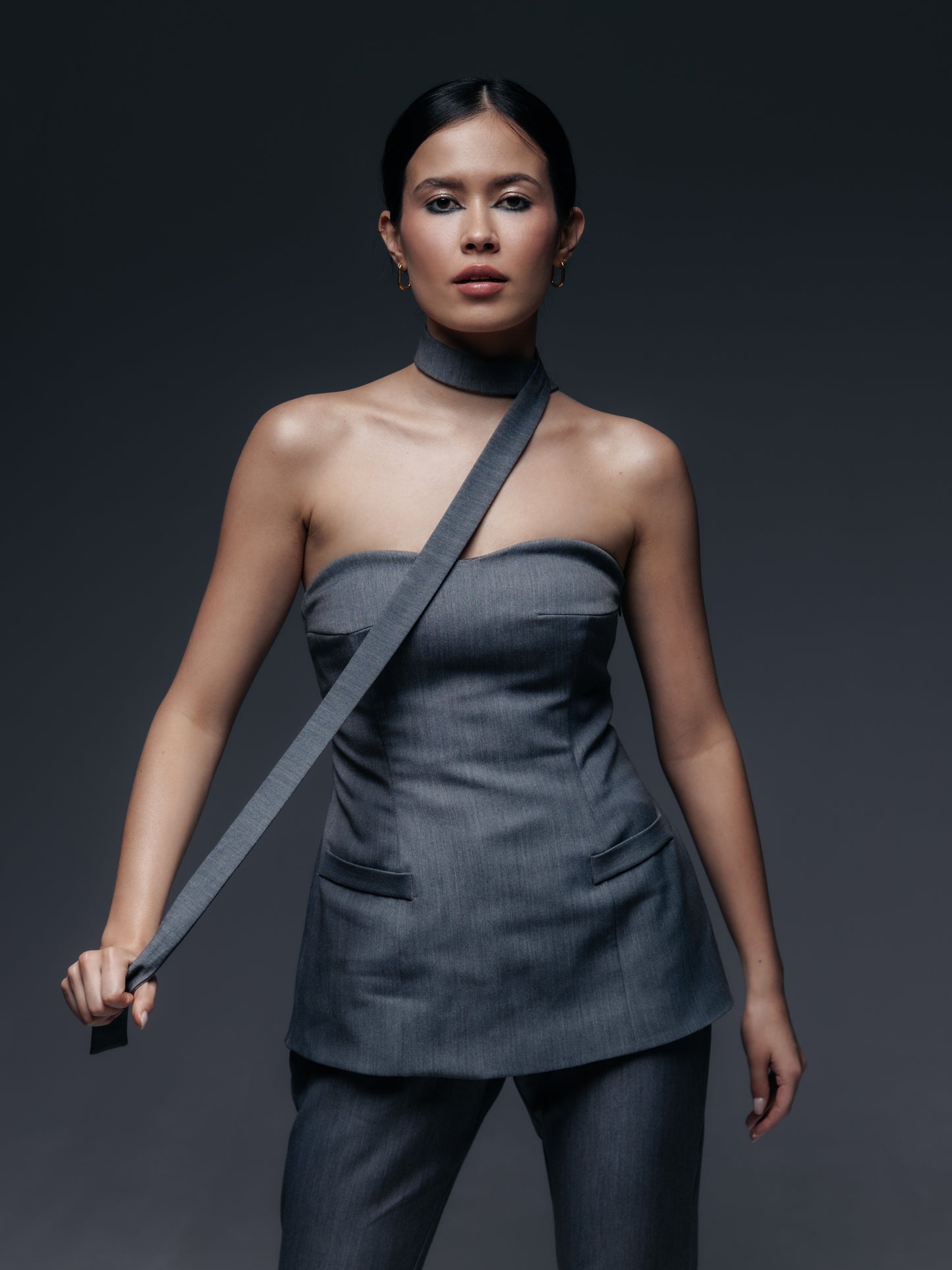 Szary gorsetowy top Mars zaprezentowany na modelce w komplecie z pasującymi spodniami Mars. Top ma fason przedłużonego gorsetu: sięga za linię talii, lekko na biodra, przypominając krótki gorsetowy żakiet bez rękawów. Wykonany z tej samej eleganckiej tkaniny garniturowej (w odcieniu grafitowej szarości) co spodnie, posiada dwie ozdobne patki imitujące kieszenie z przodu, co nawiązuje do stylu marynarki. Dekolt jest prosty, gorsetowy – top trzyma się dzięki wbudowanym fiszbinom i dopasowanemu krojowi.