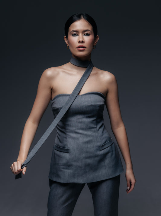 Szary gorsetowy top Mars zaprezentowany na modelce w komplecie z pasującymi spodniami Mars. Top ma fason przedłużonego gorsetu: sięga za linię talii, lekko na biodra, przypominając krótki gorsetowy żakiet bez rękawów. Wykonany z tej samej eleganckiej tkaniny garniturowej (w odcieniu grafitowej szarości) co spodnie, posiada dwie ozdobne patki imitujące kieszenie z przodu, co nawiązuje do stylu marynarki. Dekolt jest prosty, gorsetowy – top trzyma się dzięki wbudowanym fiszbinom i dopasowanemu krojowi.