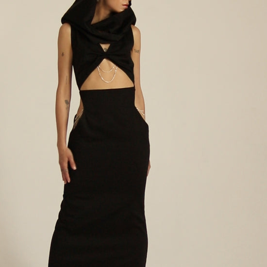 Modelka prezentuje sukienka Moon czarna, maxi z kapturem i licznymi wycięciami w talii pod biutem bogato zdobione srebrnymi łańcuszkami. Całośc ma bardzo uturystyczny klimat.