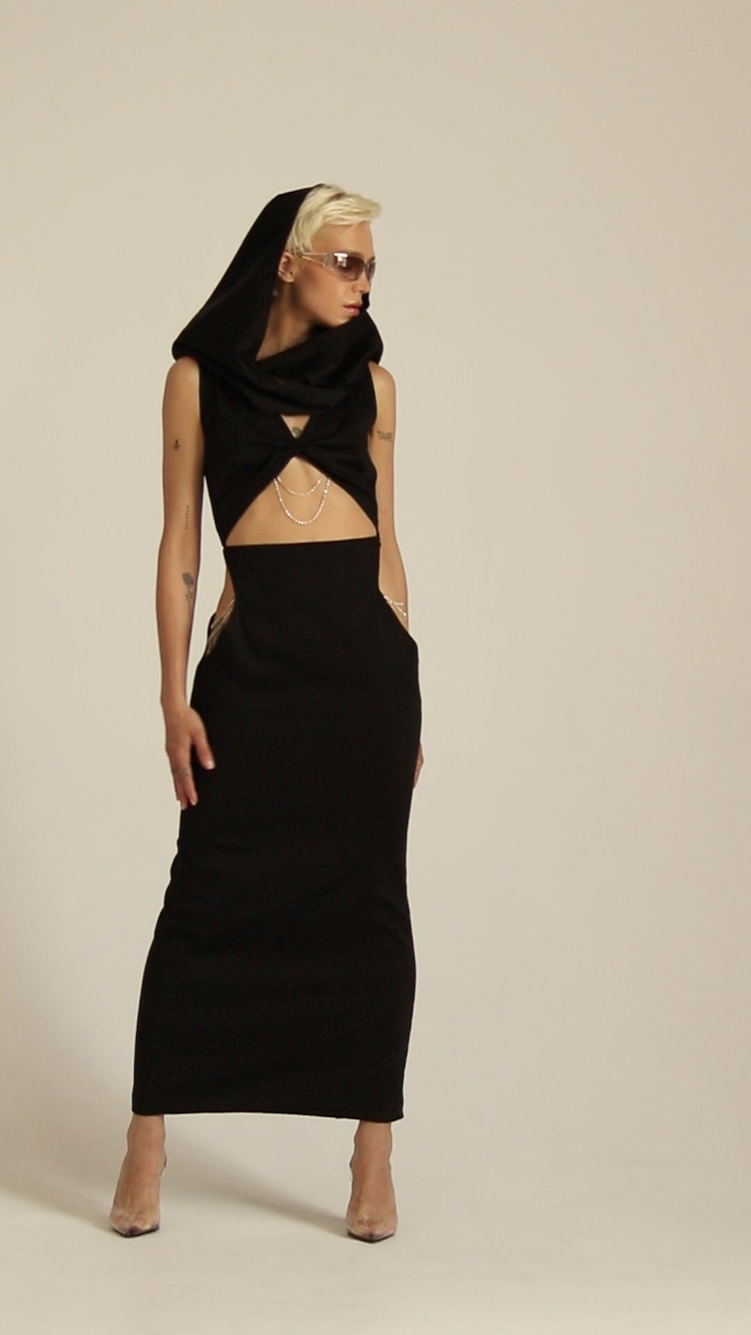 Modelka prezentuje sukienka Moon czarna, maxi z kapturem i licznymi wycięciami w talii pod biutem bogato zdobione srebrnymi łańcuszkami. Całośc ma bardzo uturystyczny klimat.