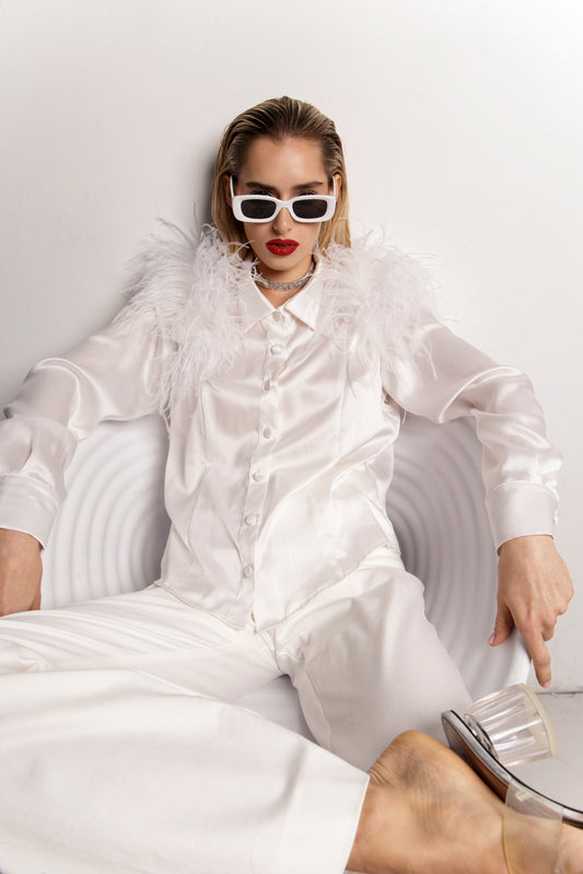 Klasyczna biała koszula jedwabna Heron z długimi rękawami, zaprezentowana w odważnej stylizacji. Ramiona koszuli zdobią bujne białe strusie pióra, tworząc efekt haute couture. Kołnierz o geometrycznym kształcie i zaszewki podkreślają elegancki, dopasowany krój. Koszula zestawiona z białymi spodniami uzyskuje monochromatyczny, ultrastylowy look na wieczorne wyjście.